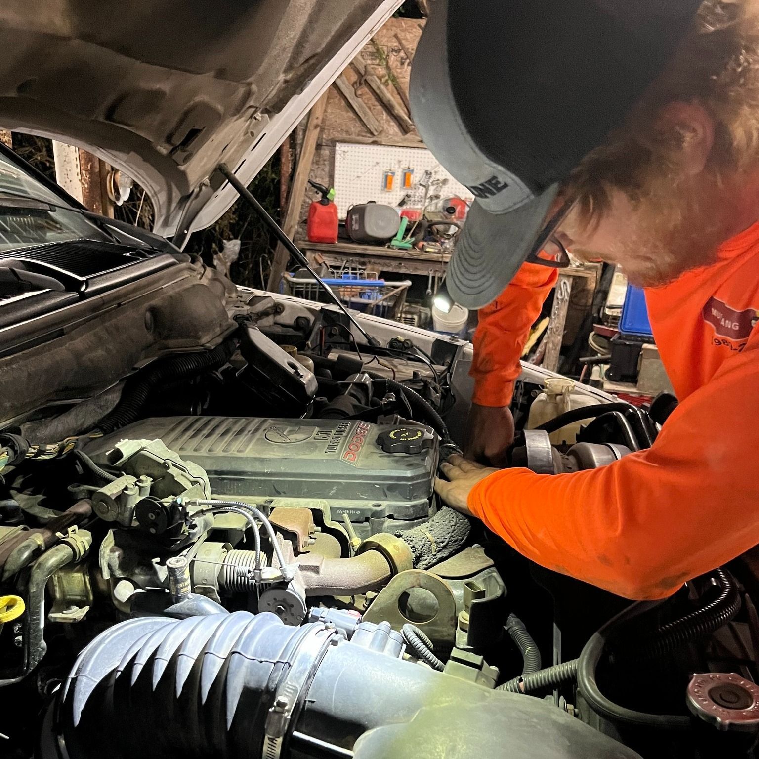 A person wearing a bright orange shirt and cap works on the engine of a vehicle in a garage.