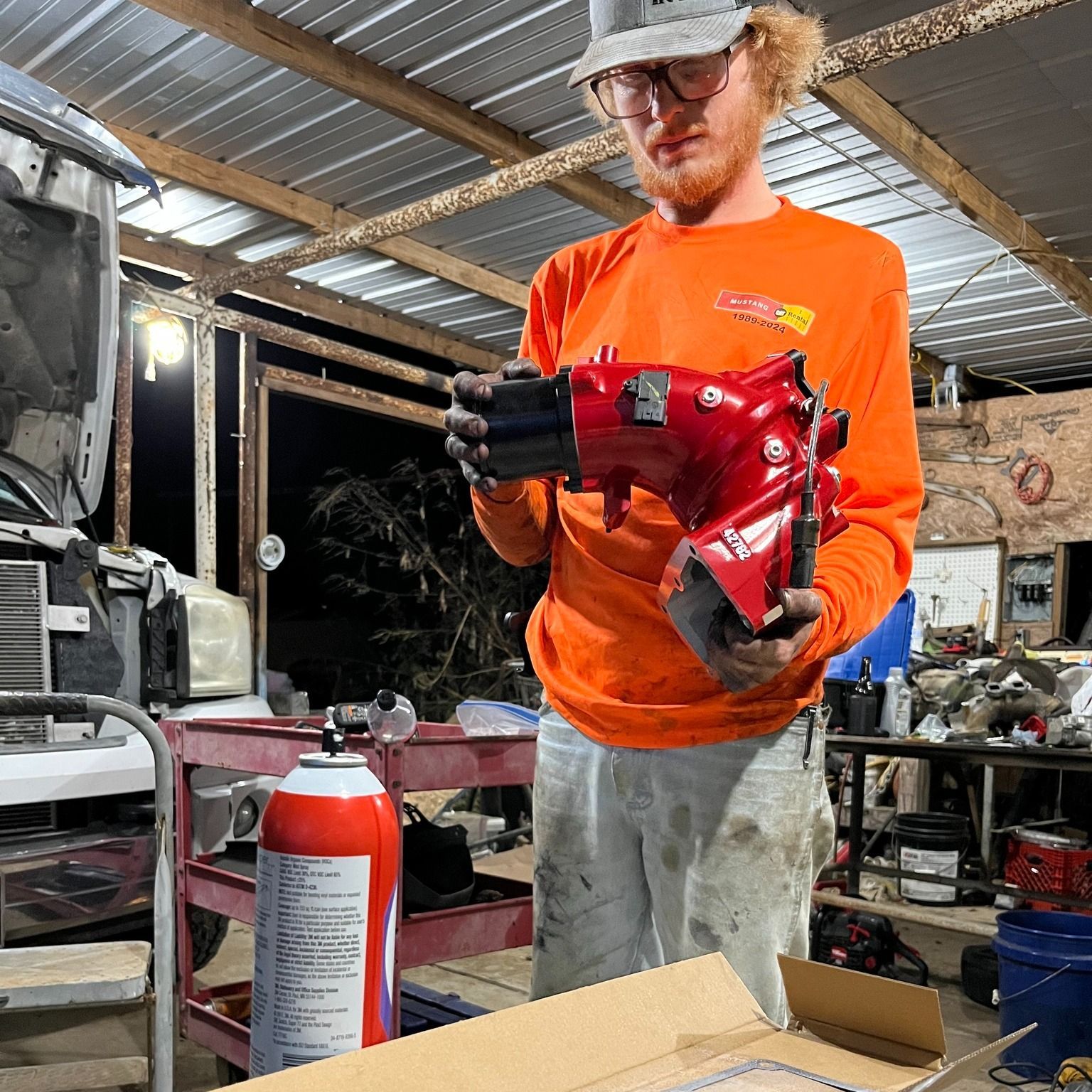 A mechanic in an orange long-sleeve shirt holds a red car part in a garage, with a fire extinguisher nearby.