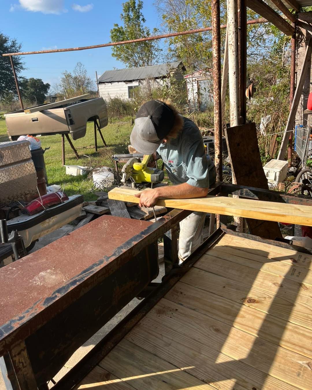A person wearing a grey cap and light-colored shirt uses a circular saw to cut a wooden plank on a trailer.