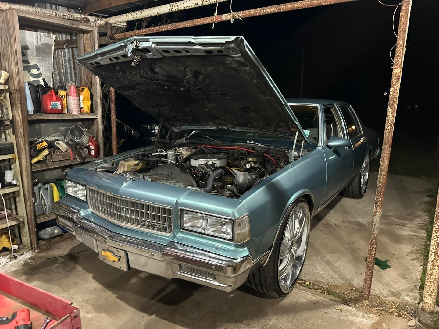 A teal Chevrolet Caprice sedan with its hood open, parked in a cluttered garage with a concrete floor.