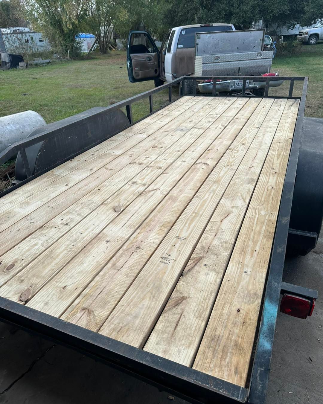 A utility trailer with a newly installed wooden plank deck parked on grass next to a white pickup truck.
