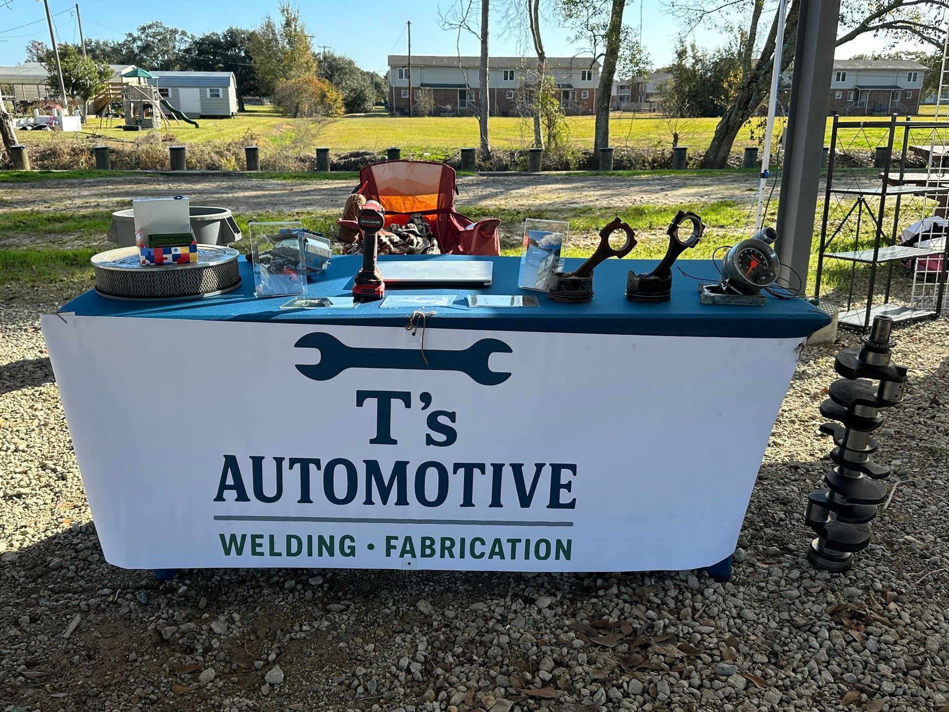 A T's Automotive table display with welding/fabrication tools and car parts on a gravel lot in front of a rural field.
