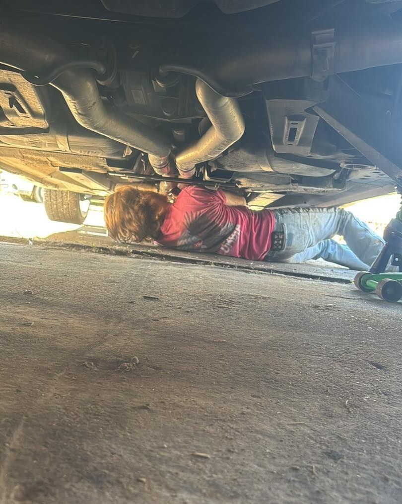 A person in a red shirt lies on the ground, working on the underside of a car raised by a green floor jack.