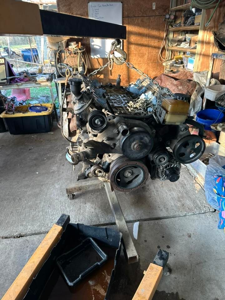 A car engine suspended by a chain hoist in a cluttered garage, with a drain pan on the concrete floor below.
