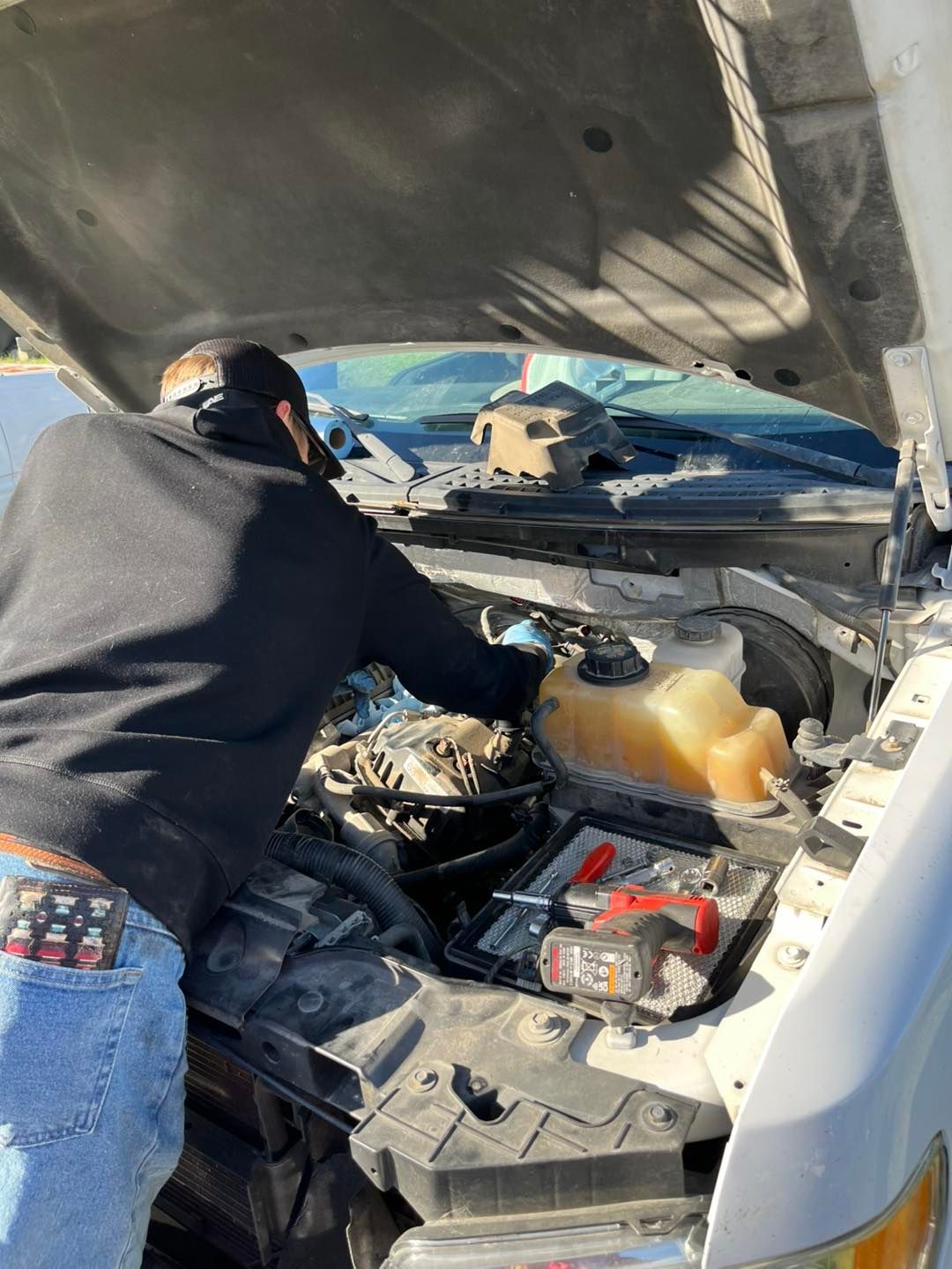 A person in a black hoodie and blue jeans leans over the open hood of a white truck to perform engine maintenance.