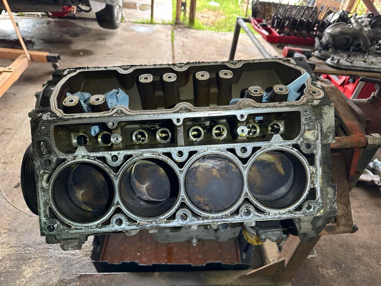 A disassembled V8 engine block sits on an engine stand in a workshop, showing open cylinder bores and internal components.