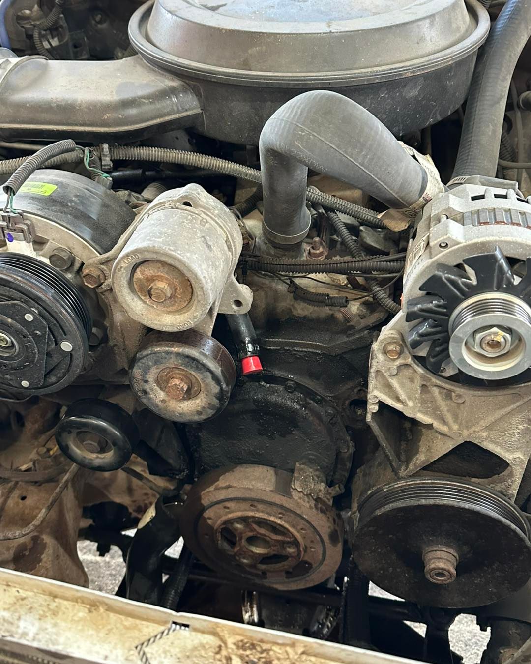 A close-up view of a dusty, used car engine with visible pulleys, hoses, an alternator, and a black air filter housing.