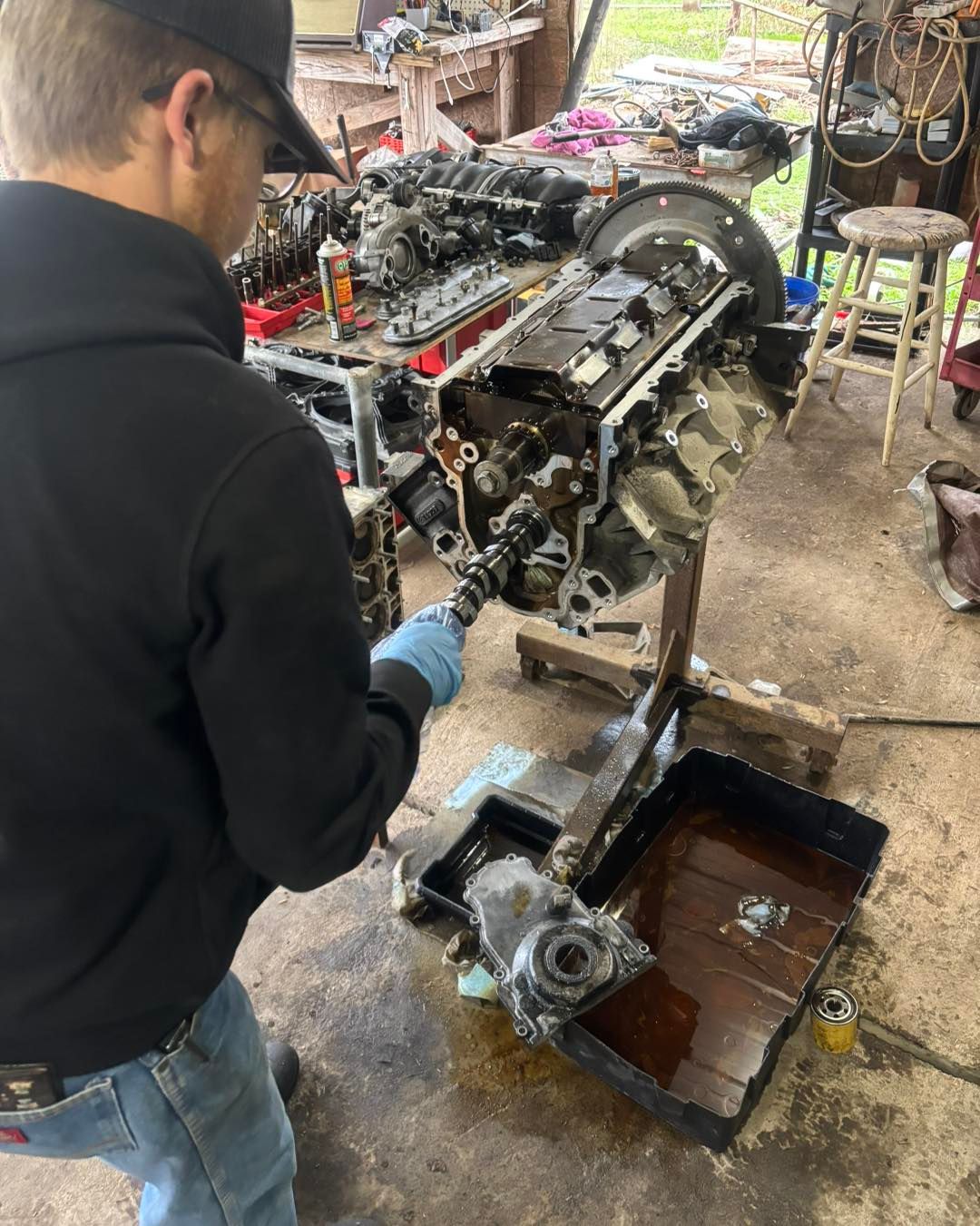 A person in a workshop wears gloves and removes a camshaft from an engine block mounted on a stand.