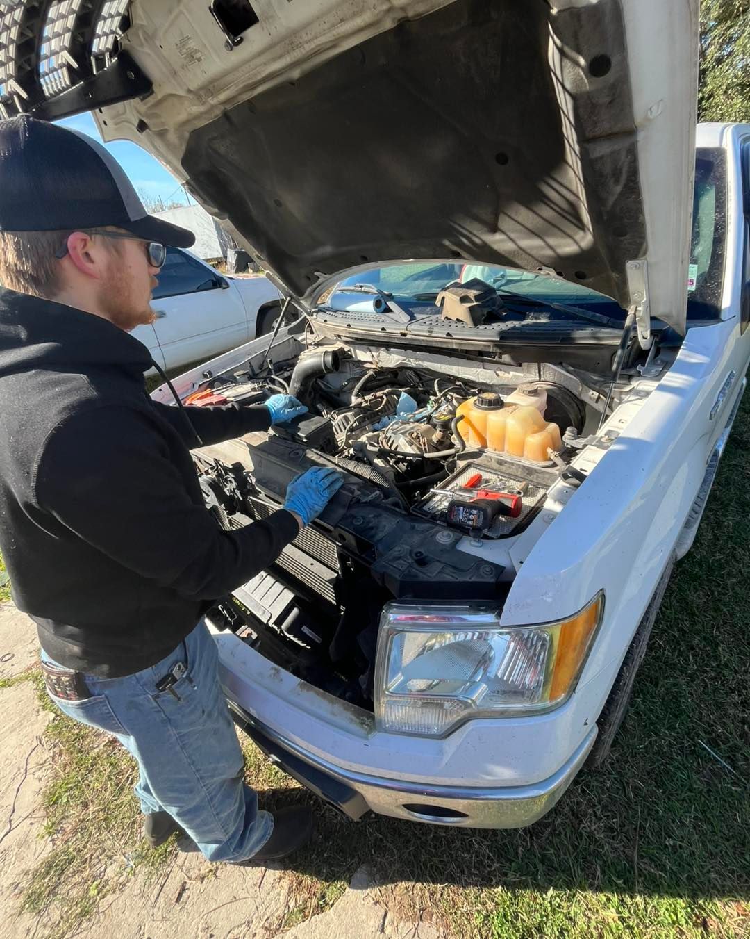 A person wearing blue gloves works on the engine of a white truck with the hood open in a sunny outdoor setting.