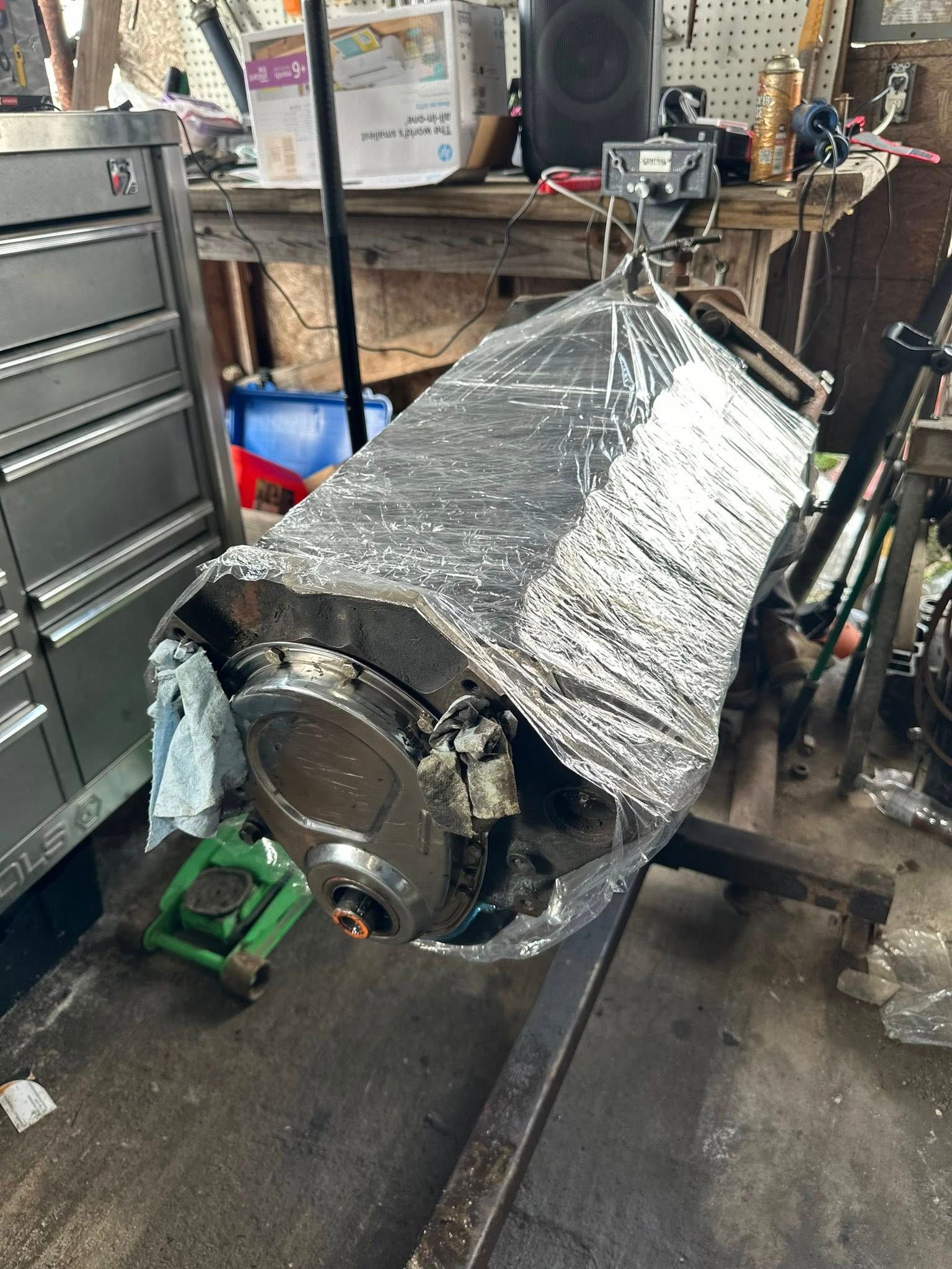 A car engine block mounted on a rotating stand in a garage, wrapped in shiny silver thermal insulation material.
