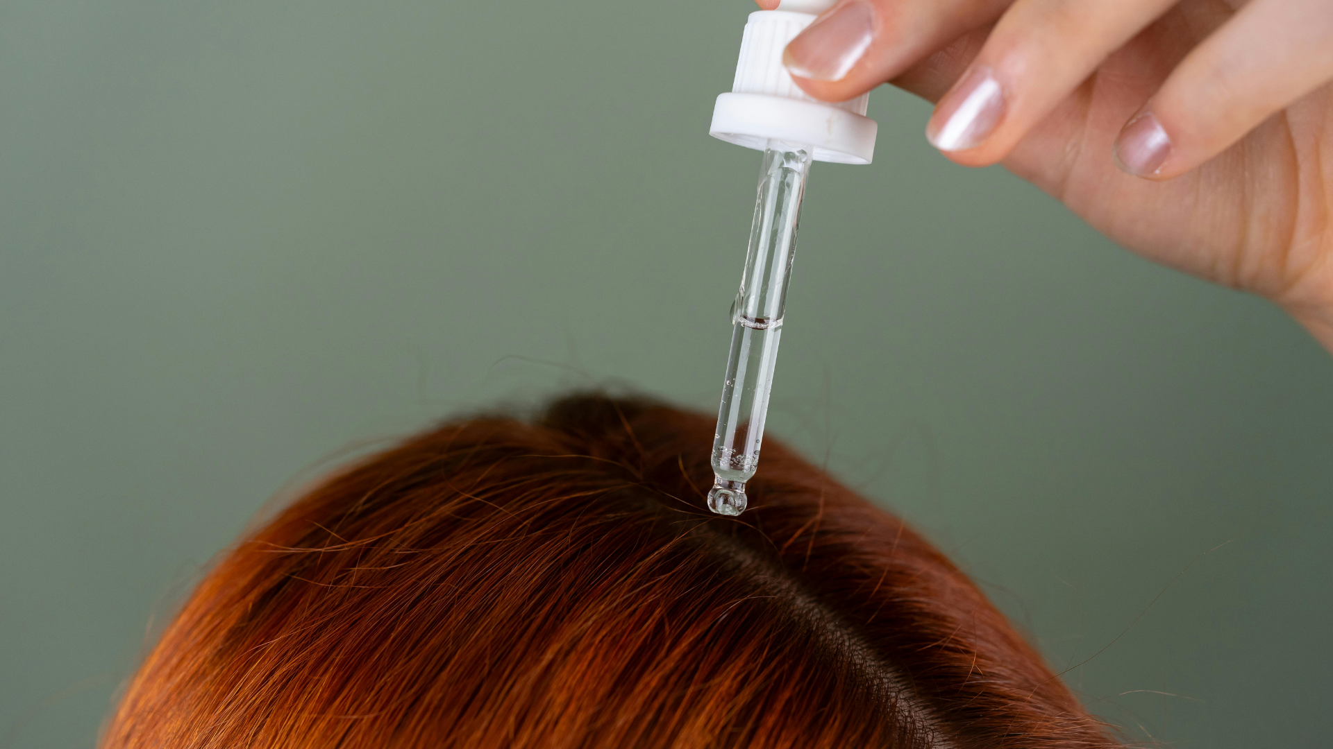 Hand using dropper to apply liquid onto a person’s reddish-brown hair parting