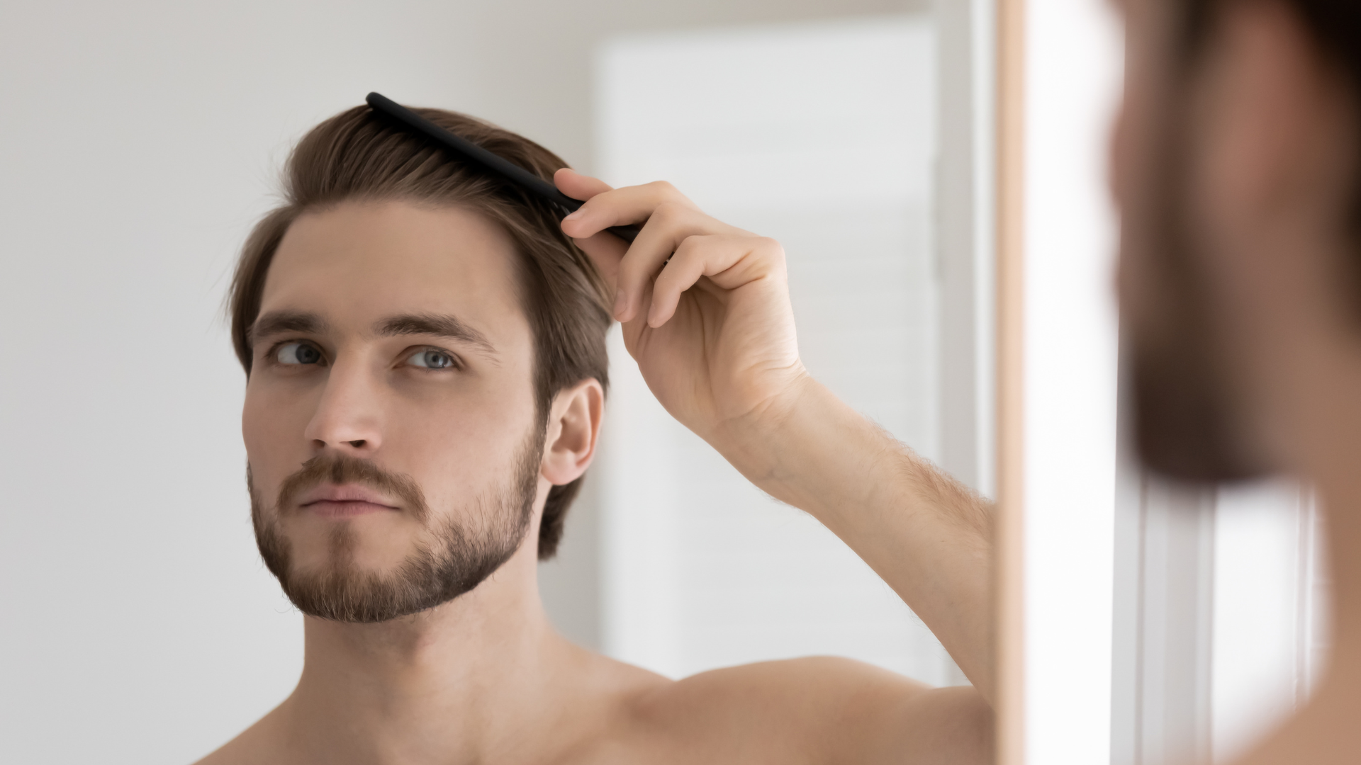 Person examining hair in a mirror, holding hair back with one hand in a bright room.