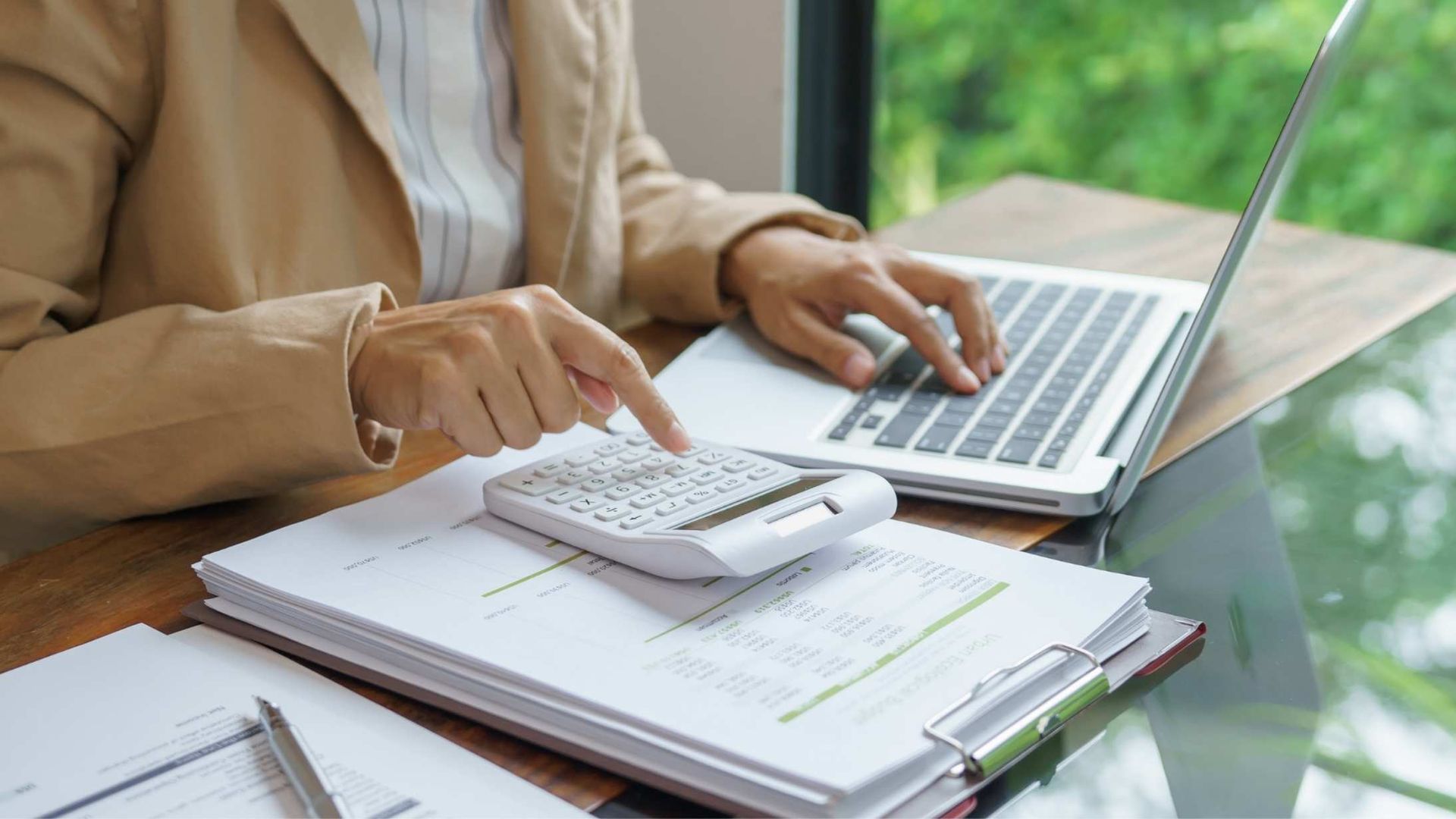 Person in tan blazer using calculator and laptop, working on documents at a desk