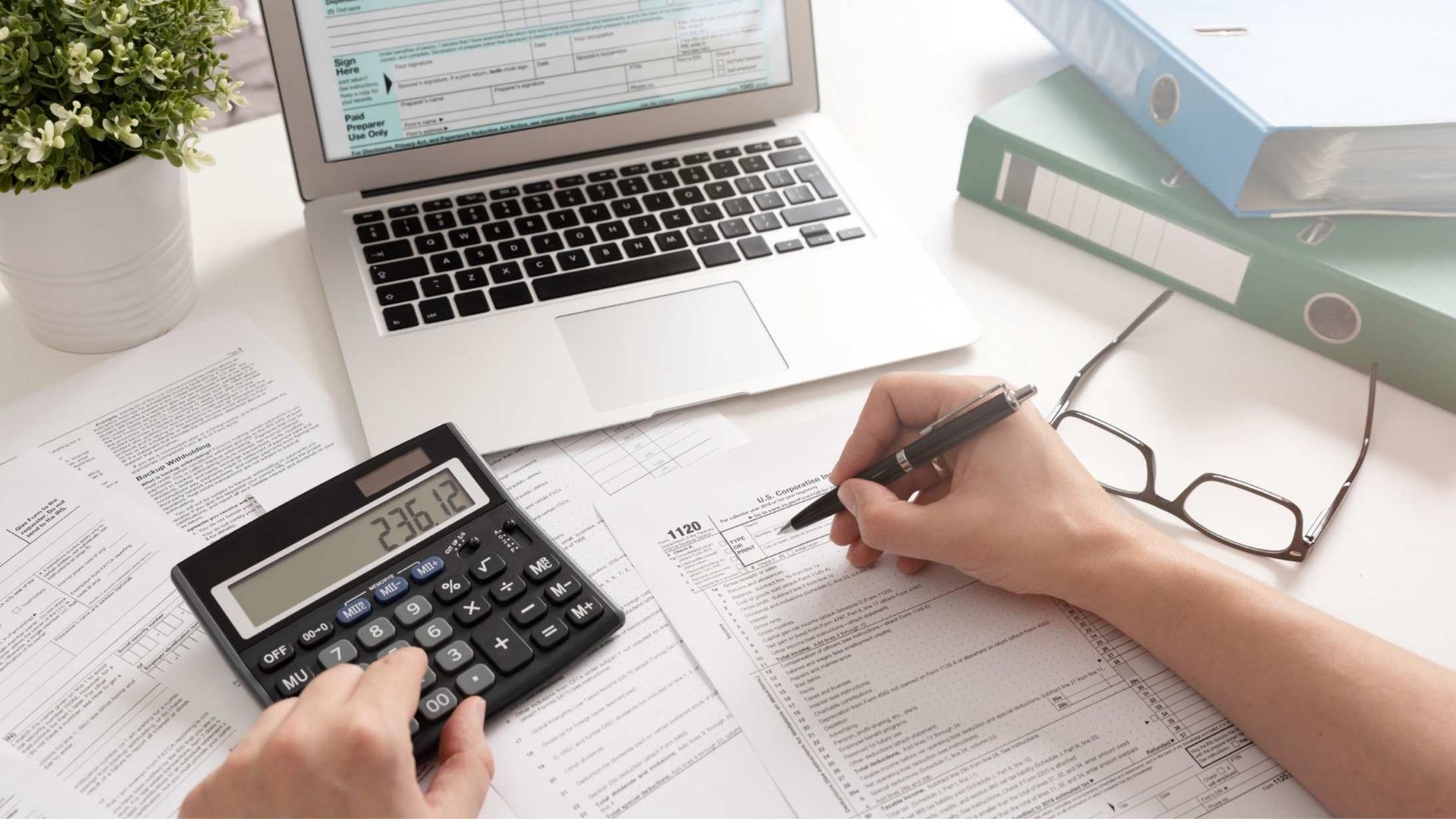 person at their laptop with a calculator in one hand and a pen with paper in the right hand