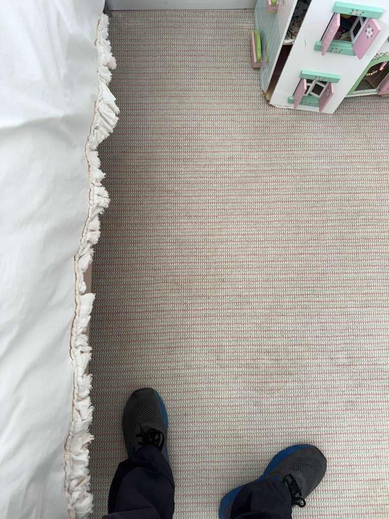Looking down at black shoes on a beige striped carpet beside a white bed and a small toy shelf.