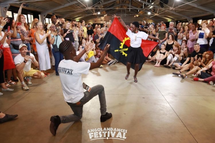 Dancers performing in a crowded indoor festival, with a red-and-black flag and seated audience watching.