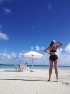 Woman in black swimsuit standing on a bright beach, looking at the ocean with a white umbrella nearby