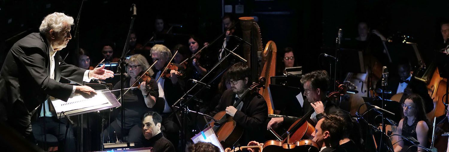 Conductor leading a dimly lit orchestra with musicians and string instruments on stage