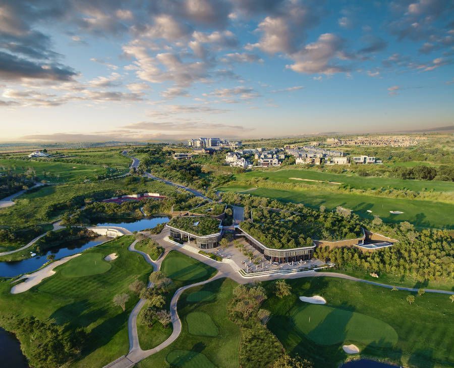 Aerial view of a green golf course with water hazards, winding paths, and buildings under a blue cloudy sky
