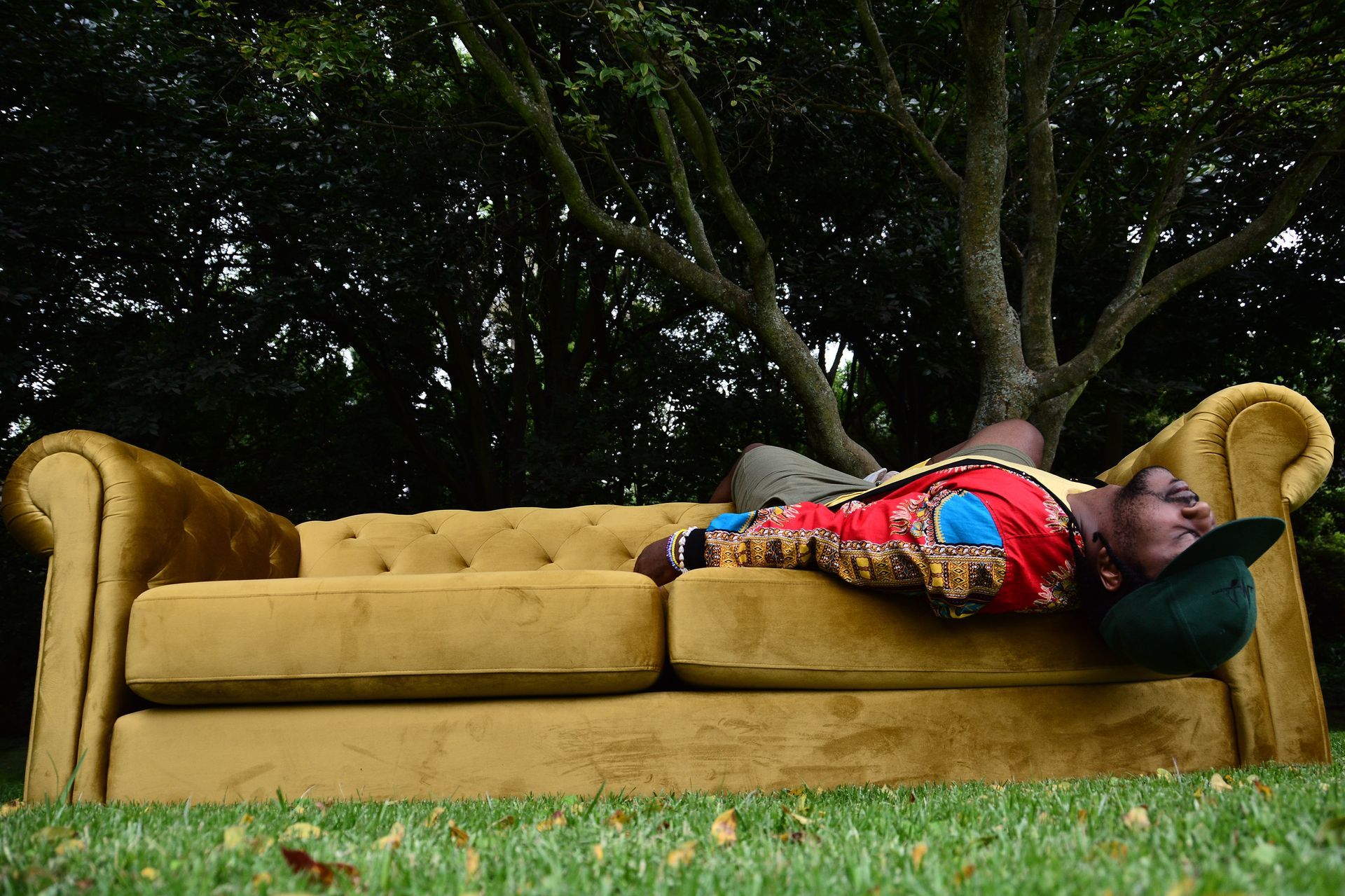 Person lying on a mustard-yellow couch outdoors on grass, with trees in the background.