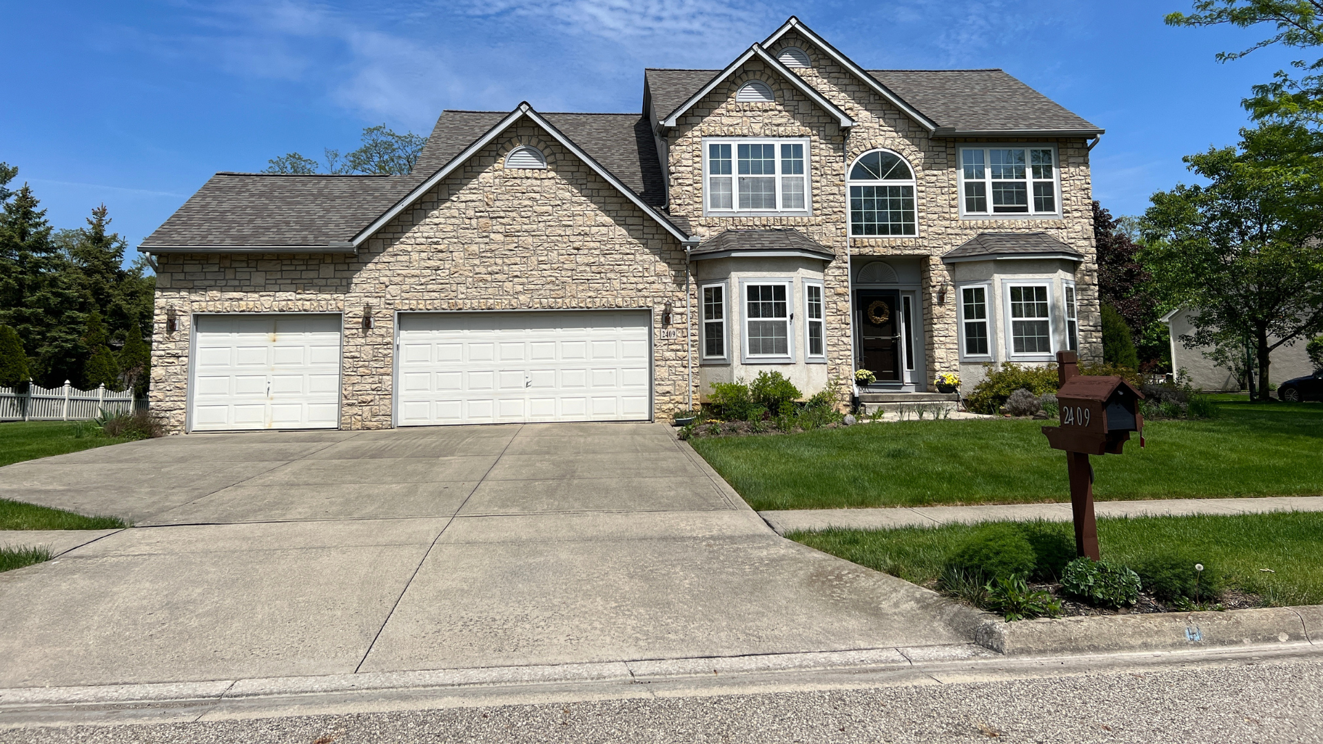 Two-story brick house with a two-car garage and driveway on a sunny day; mailbox in the foreground.