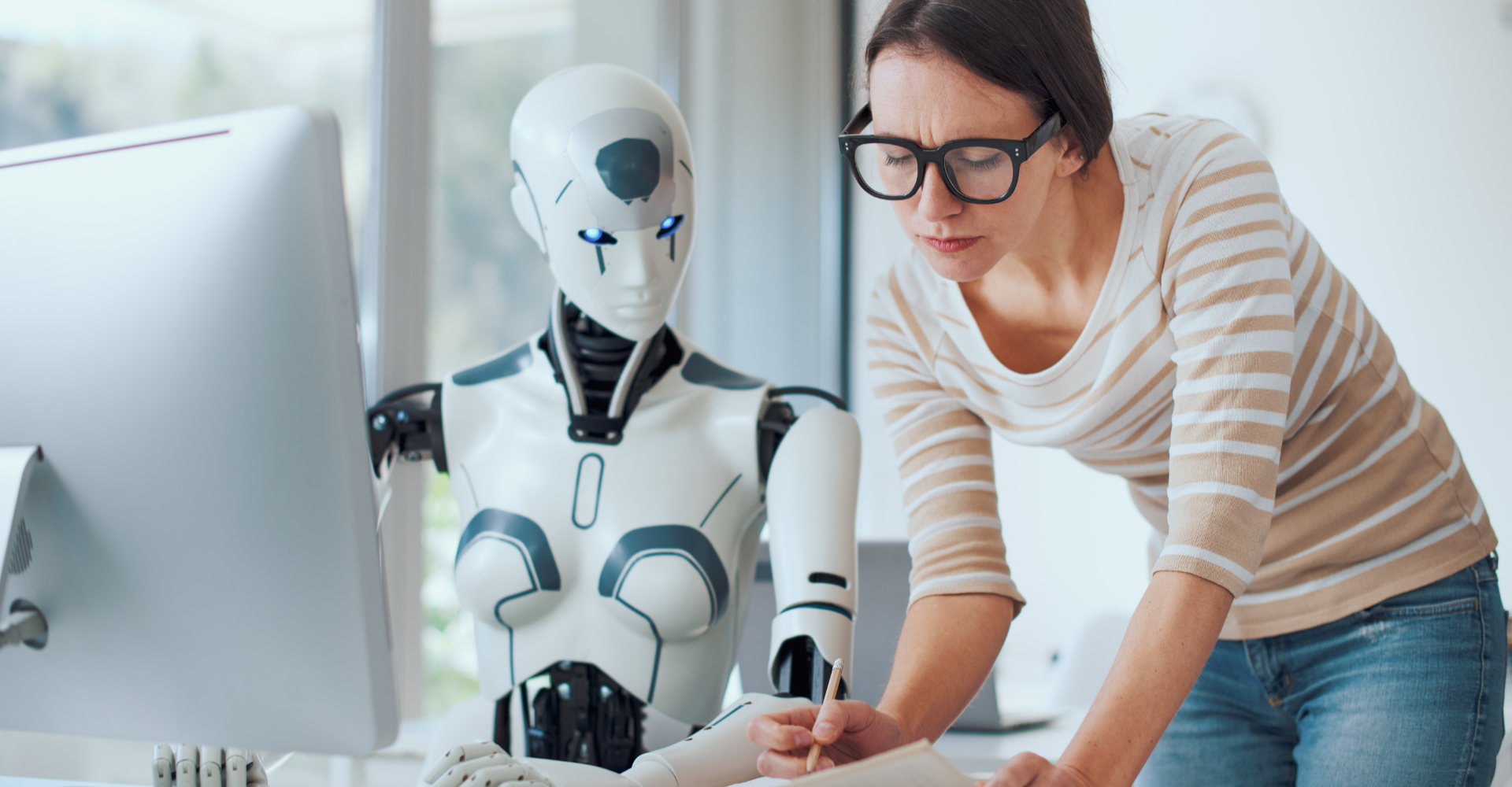 A woman leans over a desk, writing in a notebook, next to a humanoid robot with blue eyes, both focused on a computer.