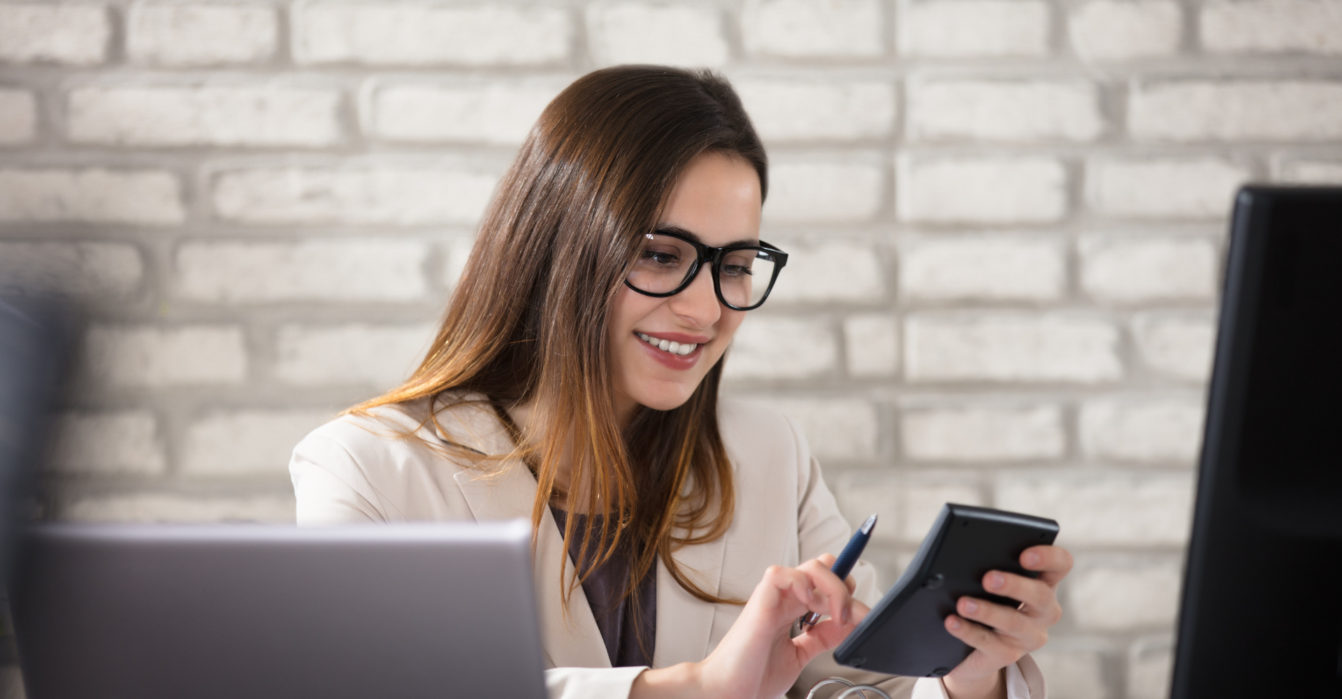 A woman with glasses, smiling and using a calculator at a desk with a laptop. She appears focused and content.