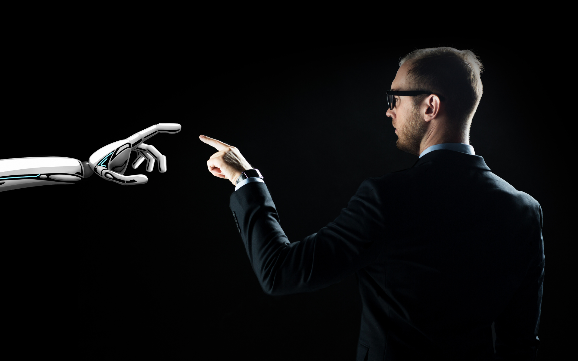 A man in a suit and glasses points towards a robotic hand against a black background.