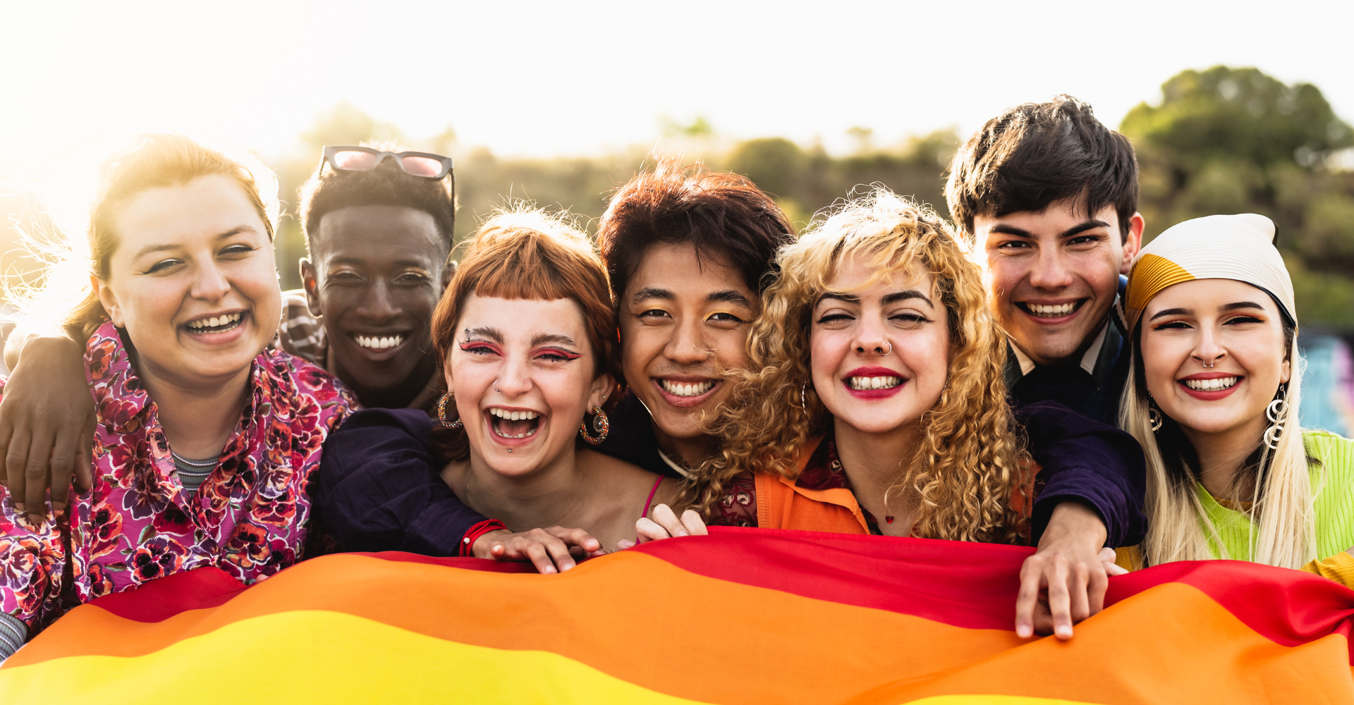A diverse group of seven smiling friends stand closely together outdoors, holding a rainbow flag. The mood is joyful.