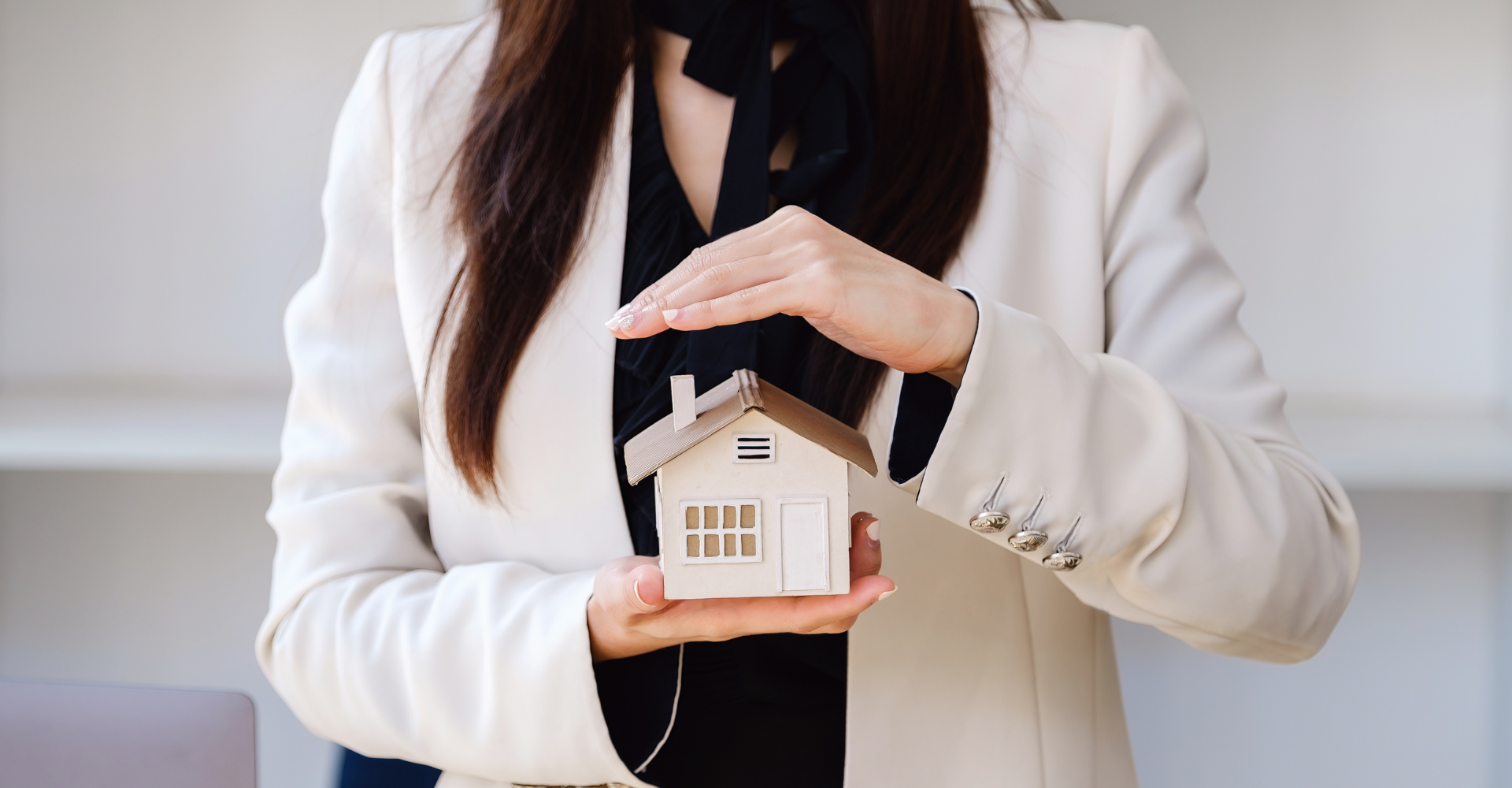 A woman holds a small wooden house model with one hand, her other hand is placed slightly above, as though sheltering it.