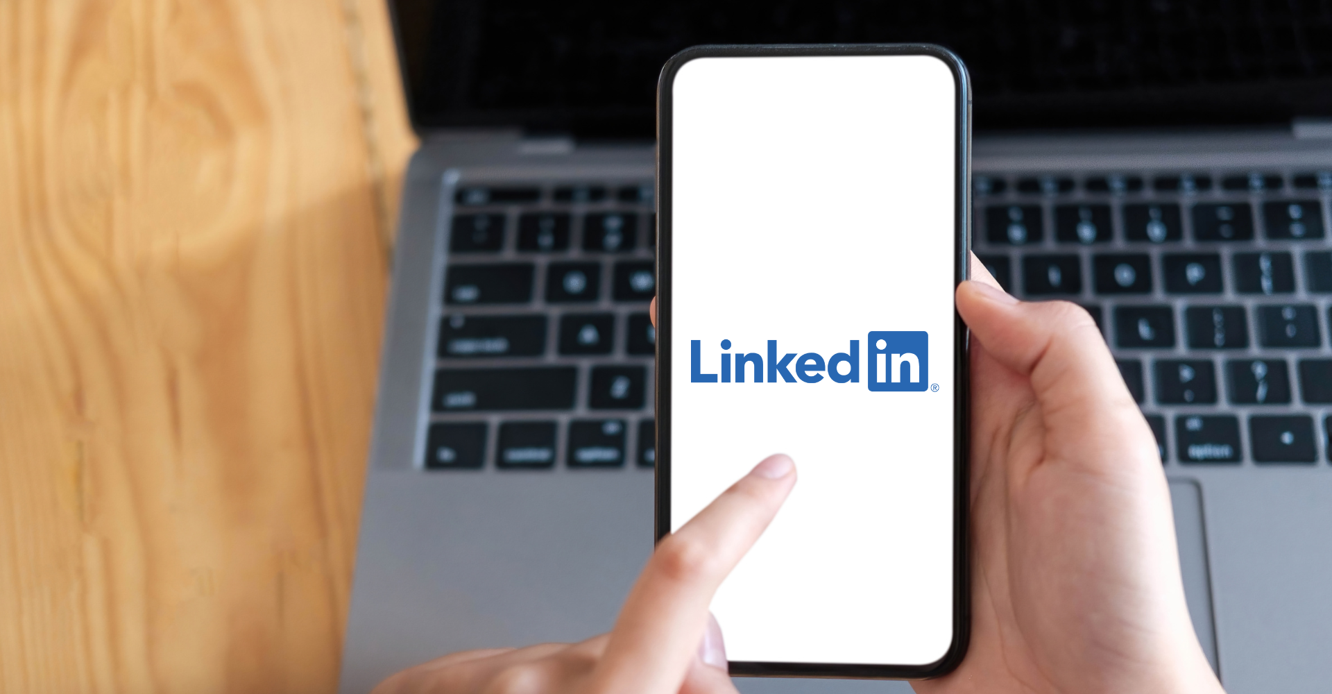 A person holds a smartphone displaying the LinkedIn logo on the screen, with a laptop on a wooden table in the background.