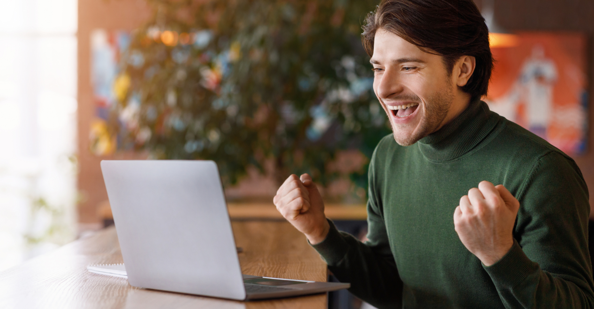 A man sits at a wooden table, celebrating in front of a laptop. Blurred background with a vibrant, cheerful ambiance.