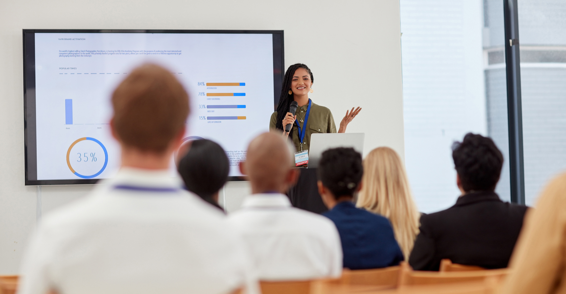 A woman presents confidently with a microphone in front of an audience. Behind her, a large screen displays charts.