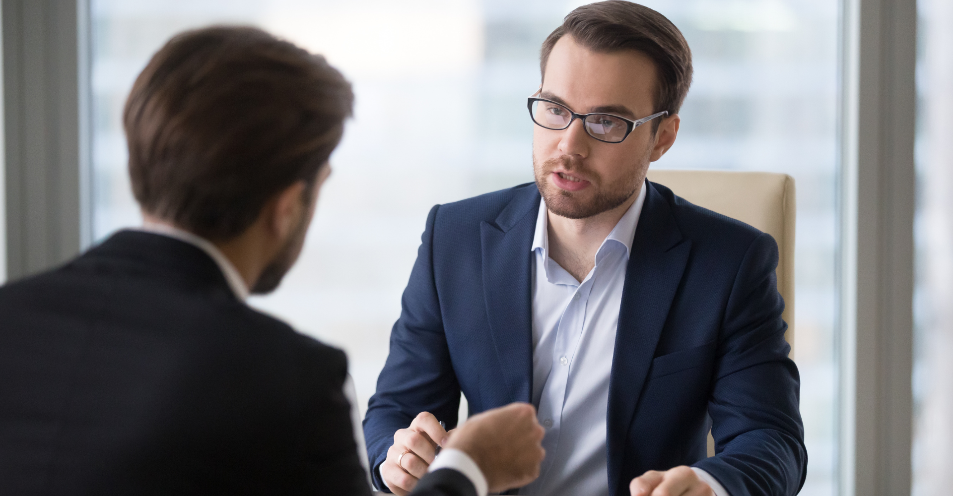 Two men in suits having a serious meeting in a bright office. One is speaking intently, while the other listens with focus. 