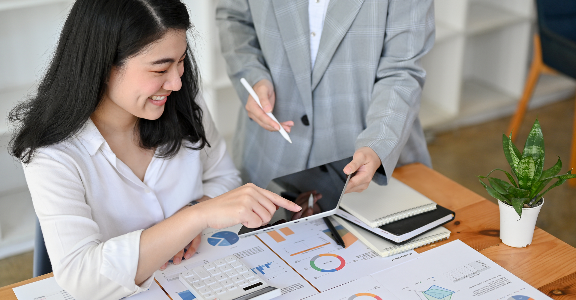 Two colleagues review charts and graphs on a table. One points at a tablet, smiling, while the other holds a pen.