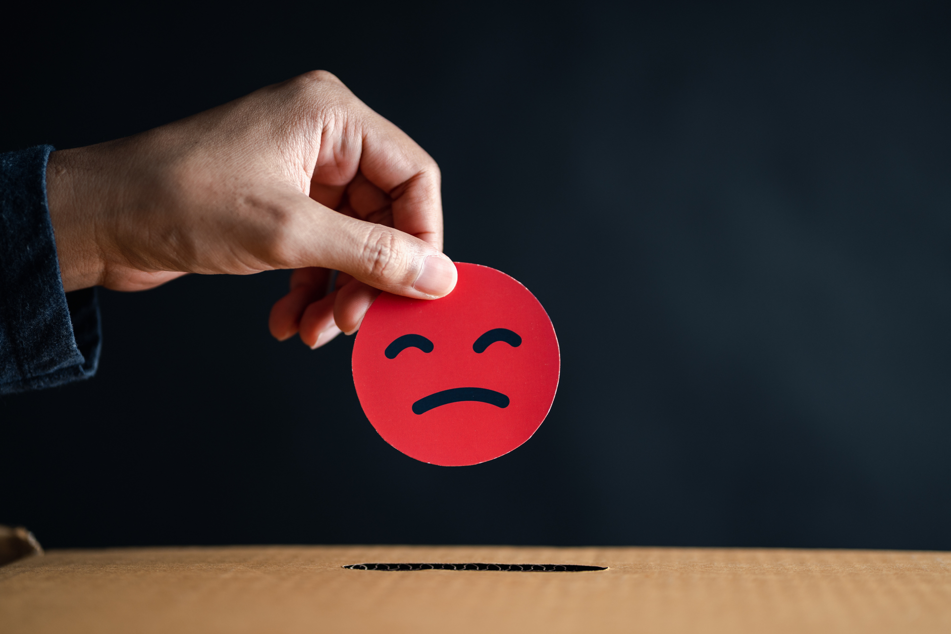 A hand holding a red paper circle with a sad face expression is poised over a cardboard box slot. The dark background