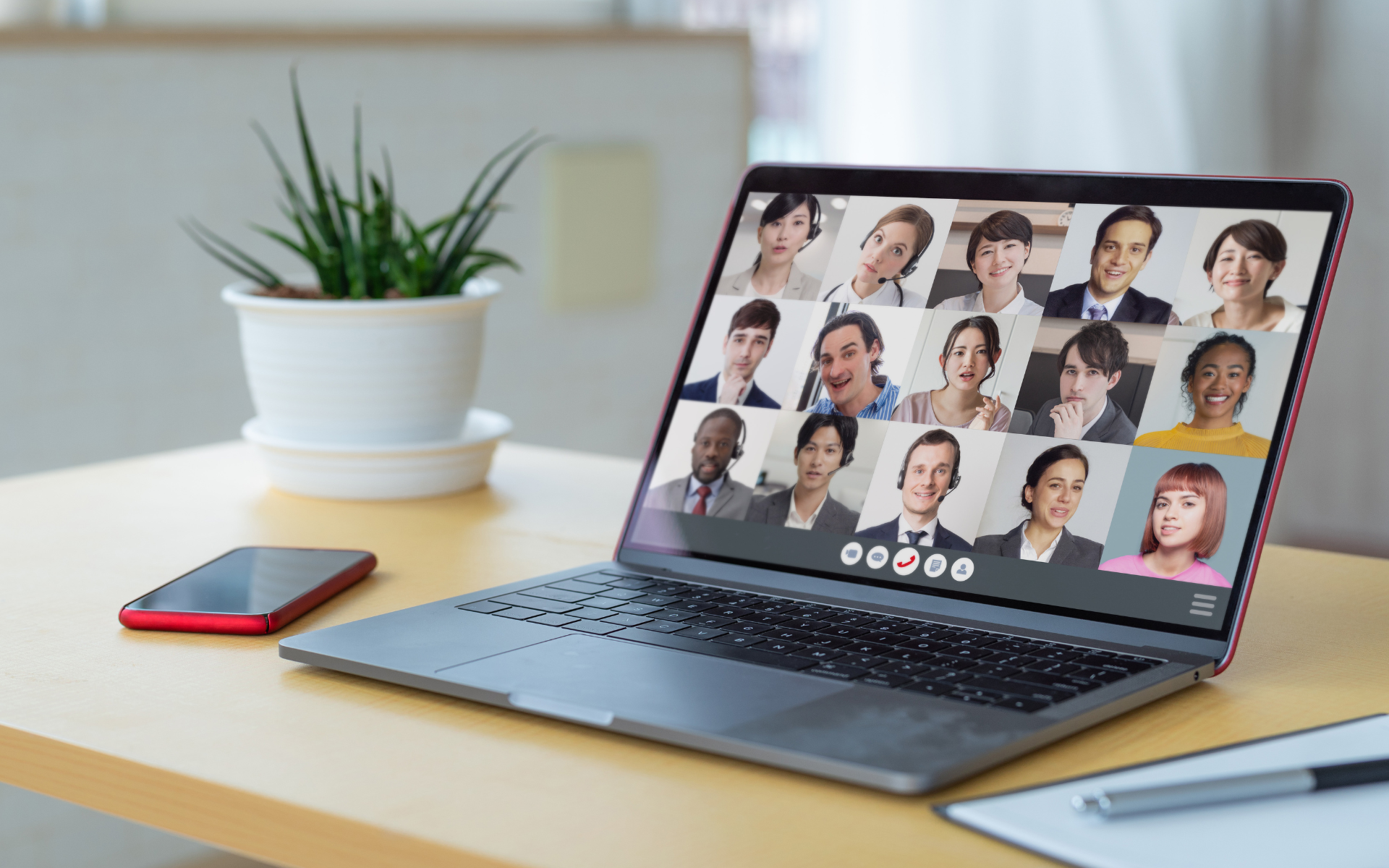 A laptop on a desk displays a grid of video call participants, each smiling. A red phone and a potted plant are nearby.