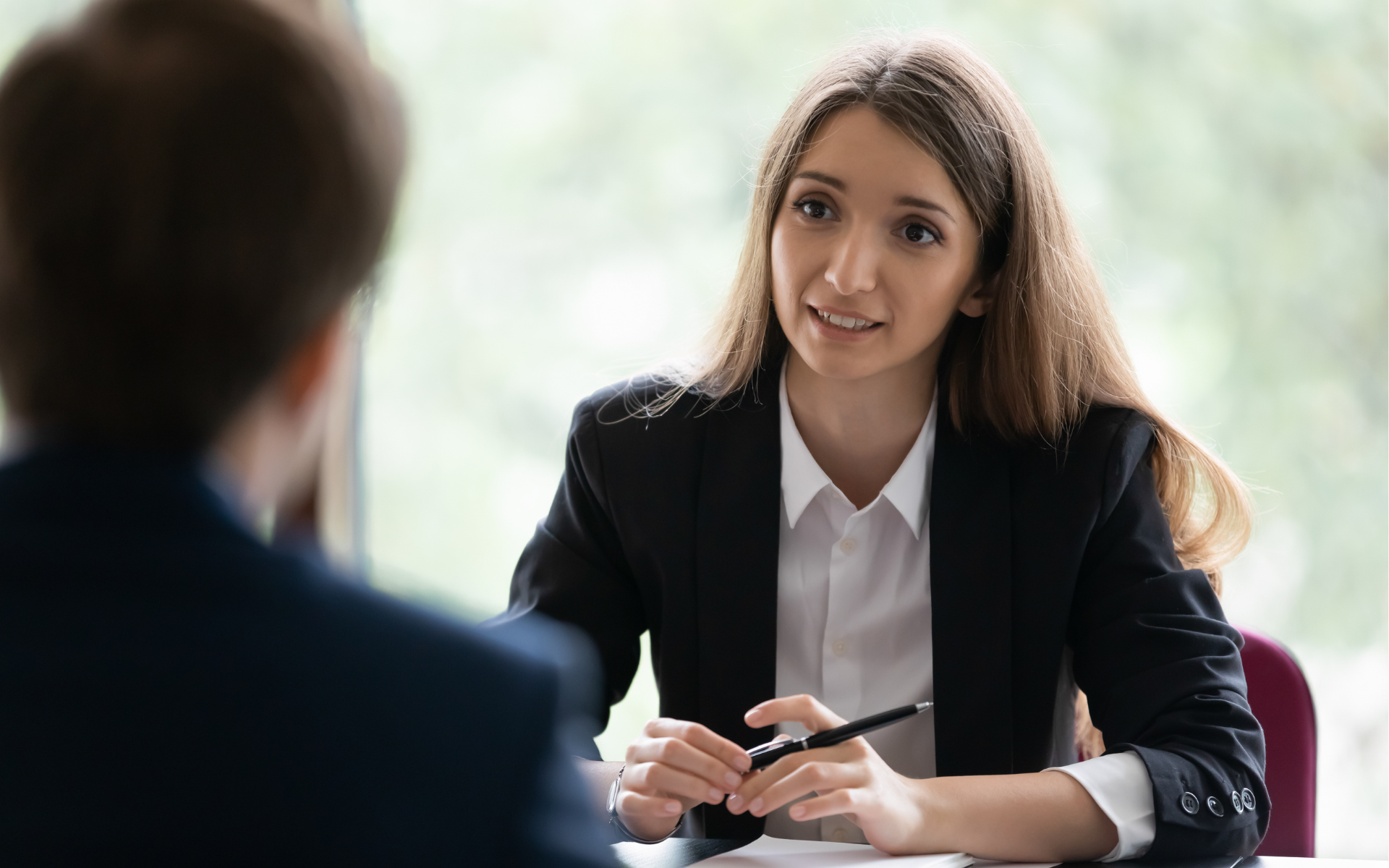Young woman in a business suit holds a pen, attentively listening and smiling. Blurred person in foreground.