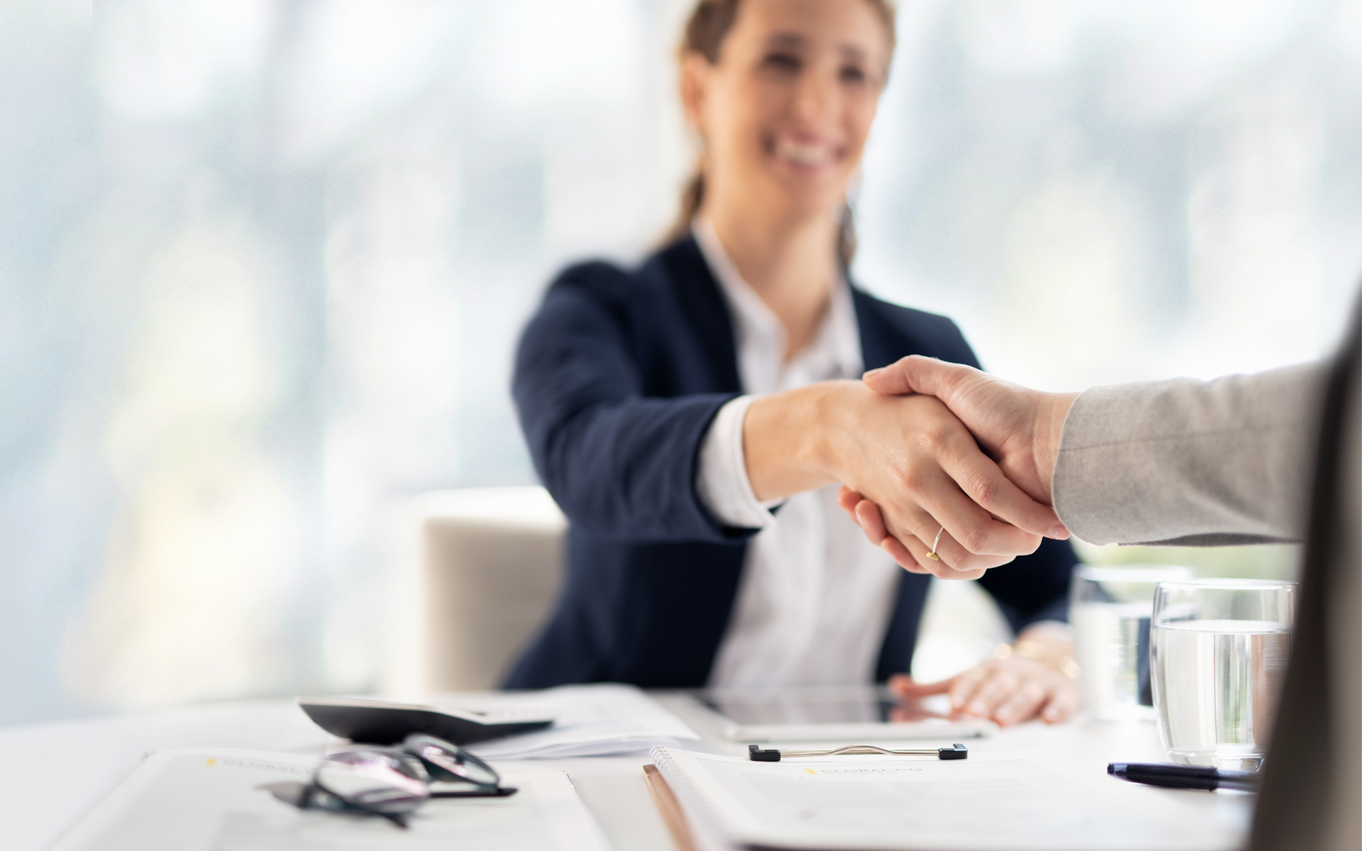 A smiling woman in a business suit shakes hands with a person across a desk, signifying a successful agreement.