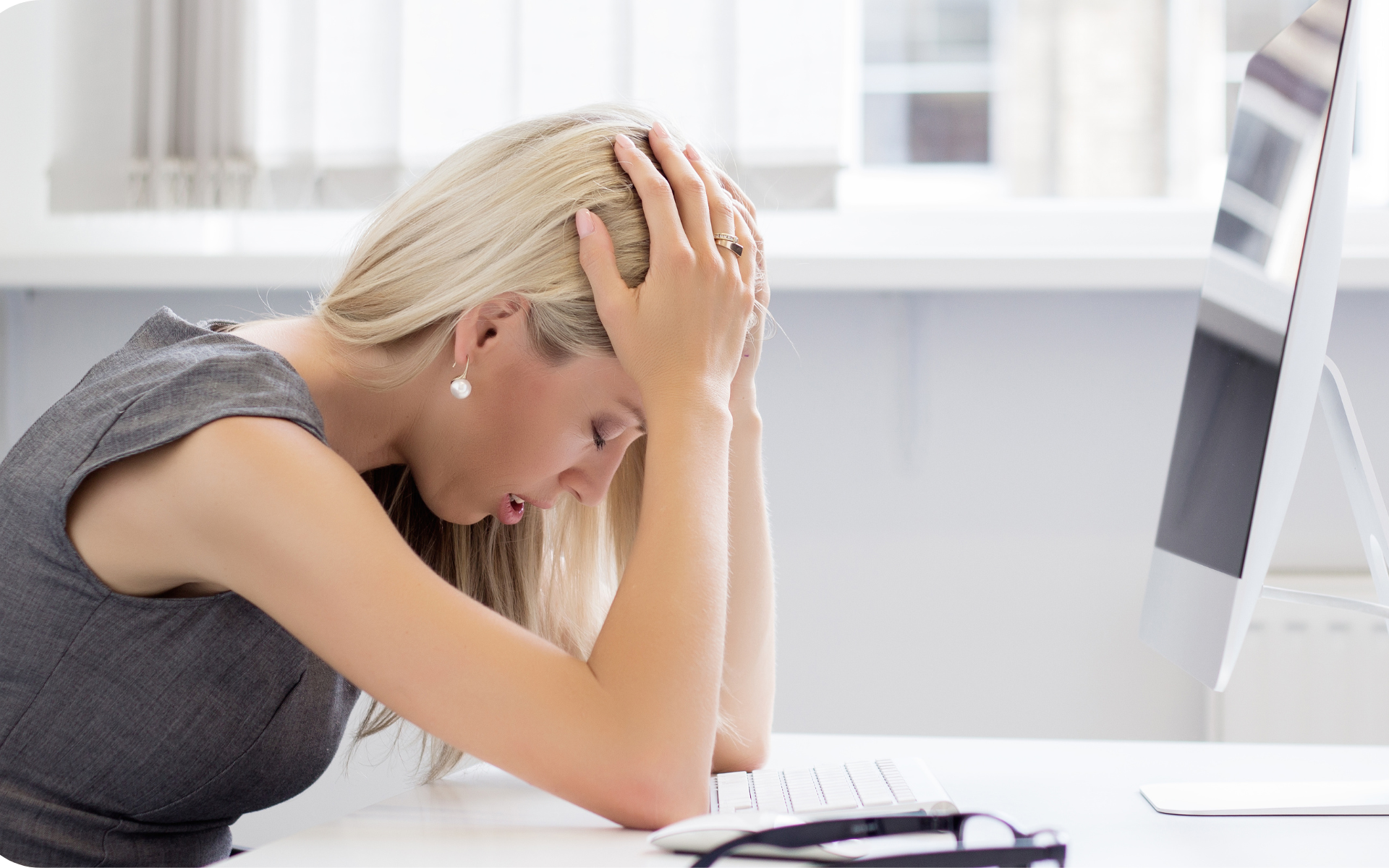 A woman sits at a desk, head in hands, appearing stressed. She is in front of a computer, conveying a feeling of frustration.