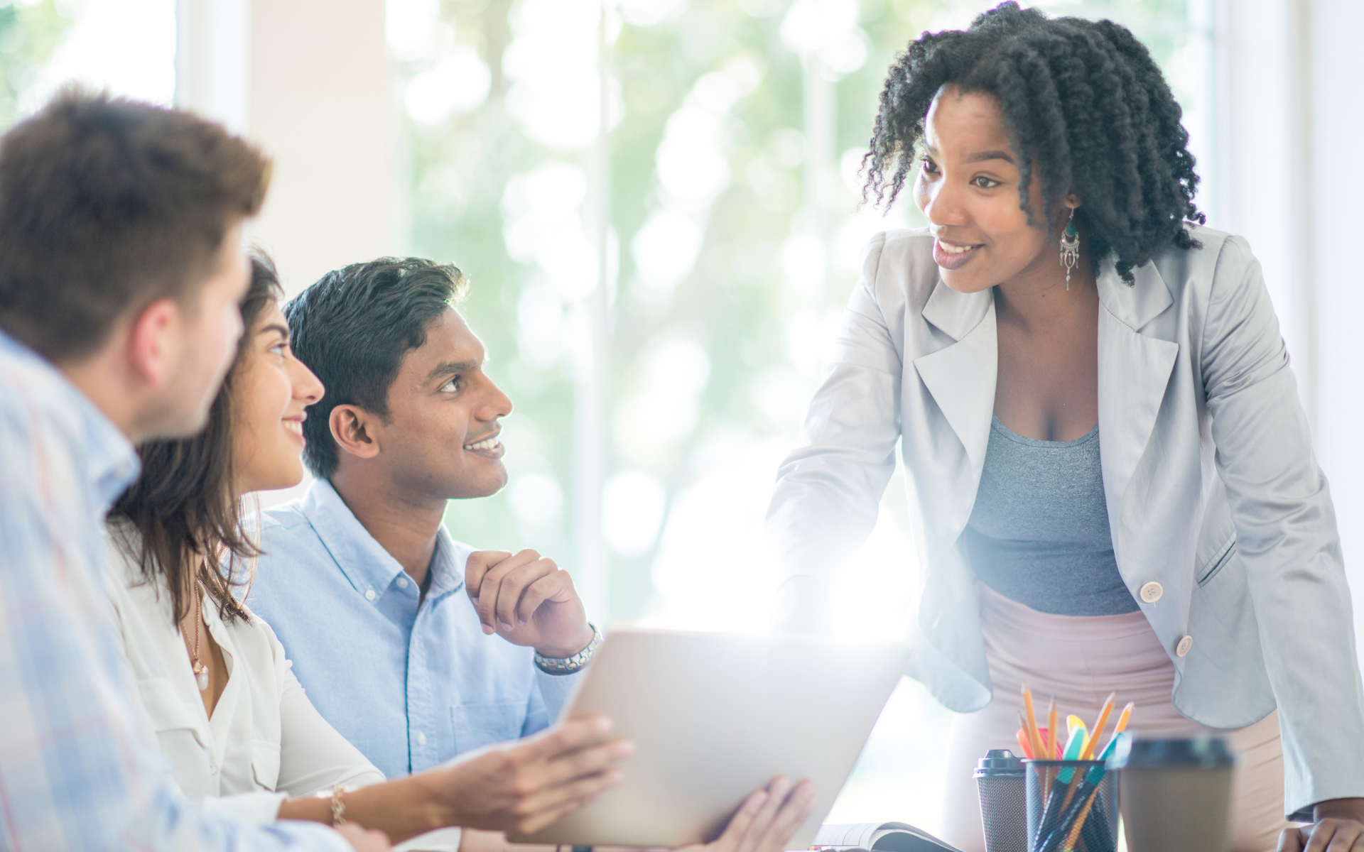 A confident woman engages three colleagues with a tablet in a bright office setting, conveying teamwork and collaboration.