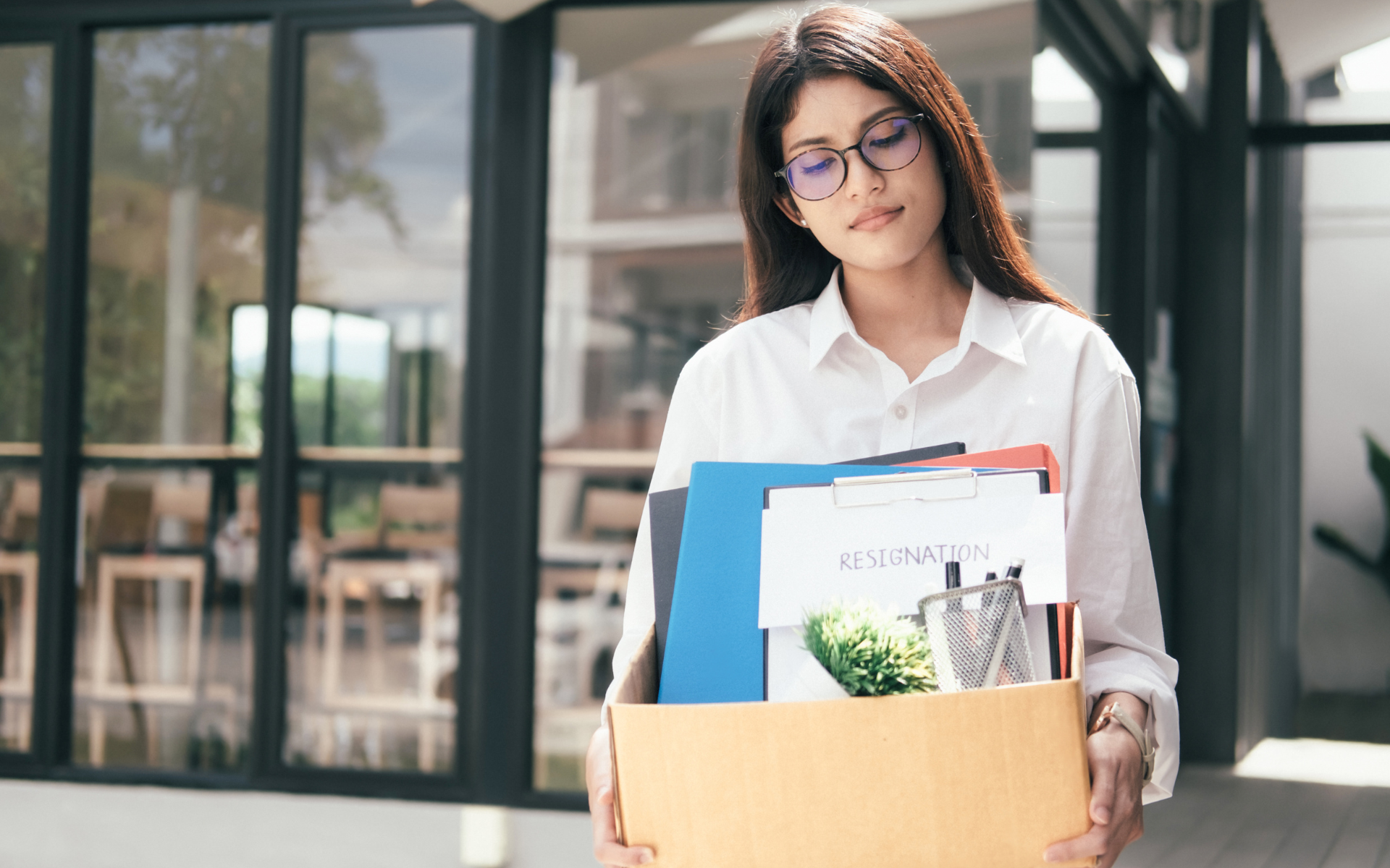 A woman wearing glasses and a white shirt holds a box with office items, including a plant and a 