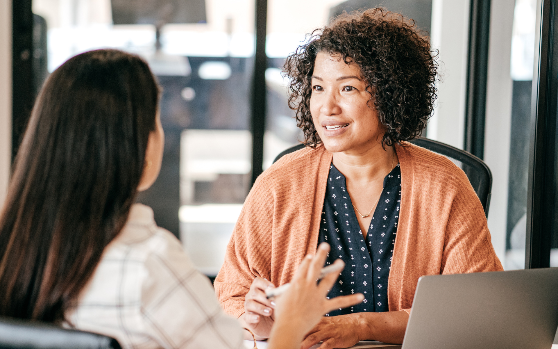 Two women engaged in a conversation at an office table. One woman smiles warmly. A laptop is open nearby.