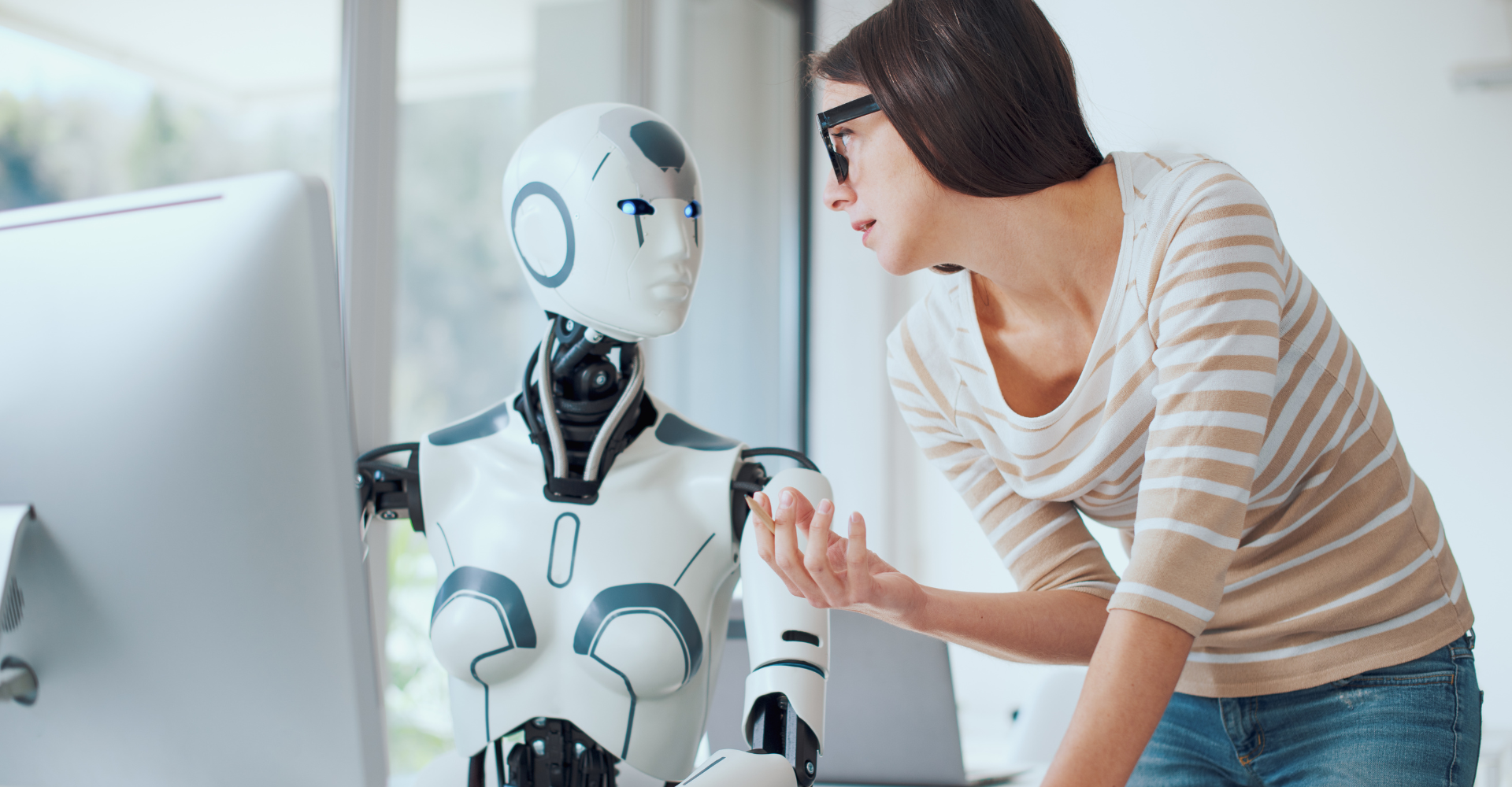 A woman in glasses is engaging with a humanoid robot in a bright room. The robot is white and futuristic.