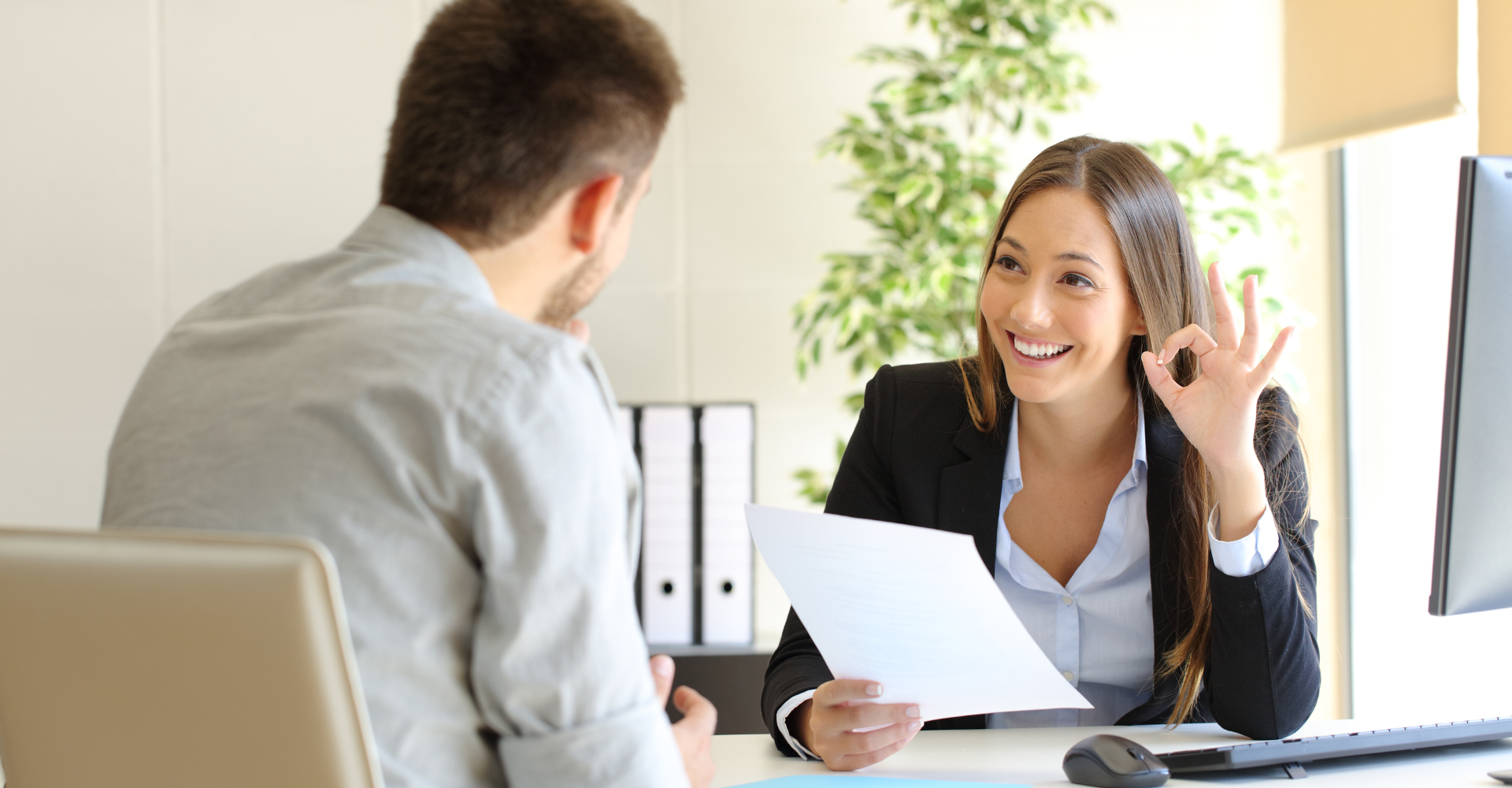 Woman smiles, holding a paper and giving an OK gesture during an office meeting. A man sits opposite, listening attentively.