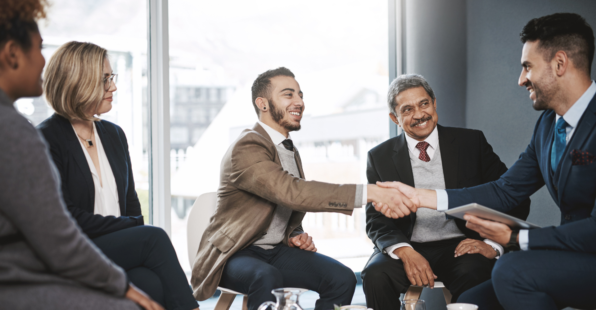 A diverse group of professionals sit in a well-lit office, two men in suits shaking hands, smiling.