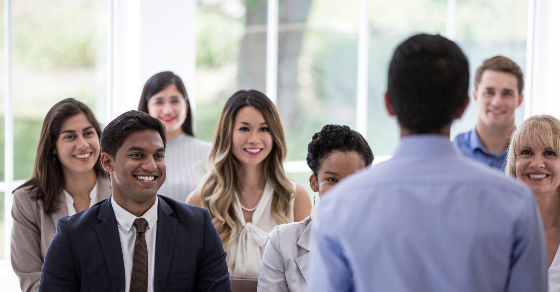 A diverse group of eight people sitting smiling and attentive. They face a speaker in focus from behind.