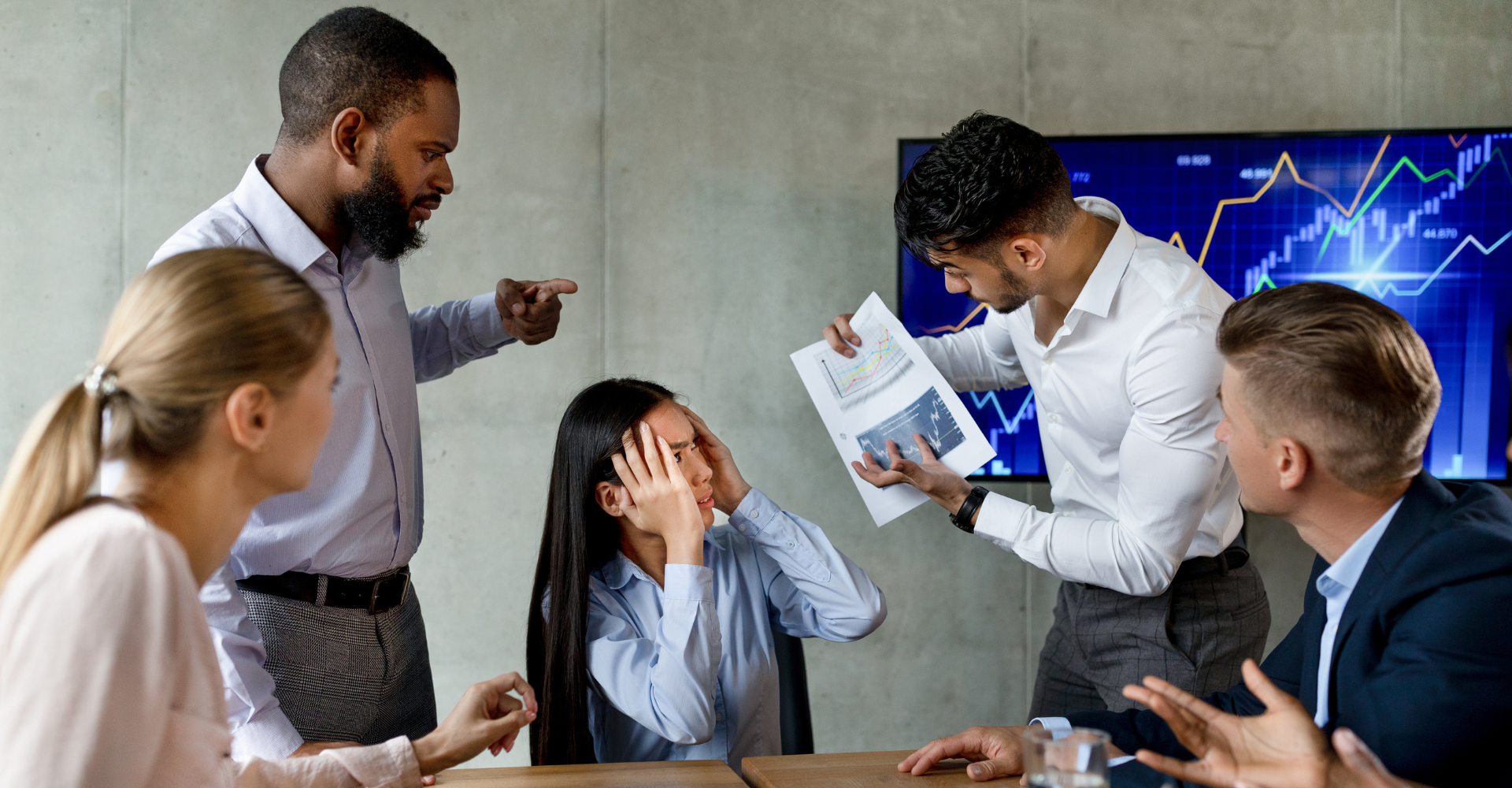 A tense business meeting with five people. A man points aggressively while another shows a distressed woman a graph.