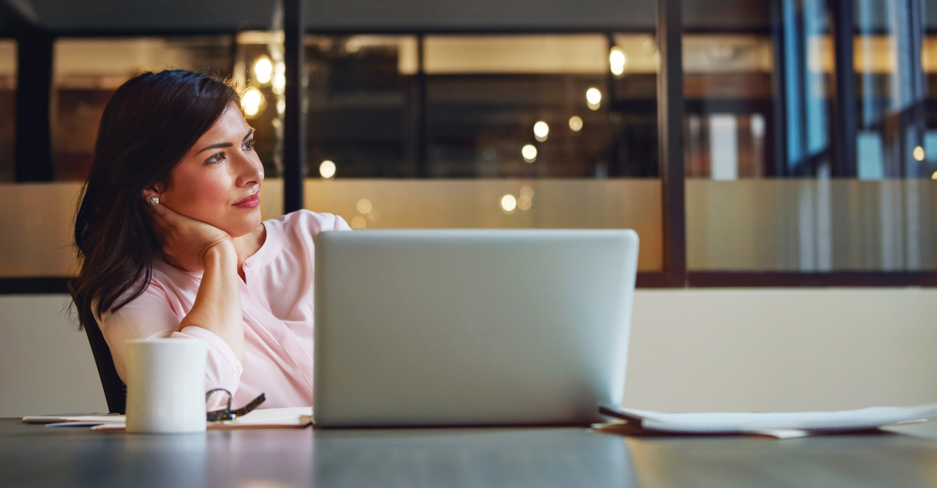 A woman in a pink blouse sits at a desk, gazing thoughtfully. A laptop, papers, and a coffee cup are on the table.