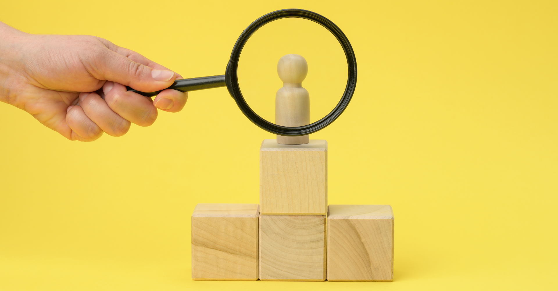 A wooden figure stands on stacked blocks against a yellow background, as a hand holds a magnifying glass above it.